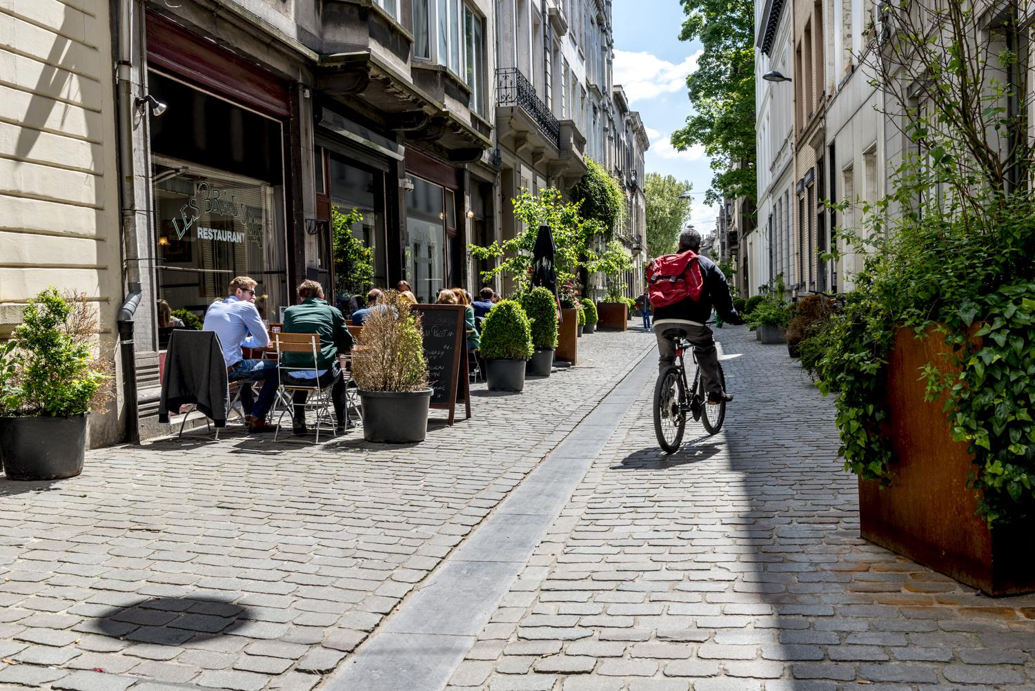 Cycliste se déplace Rue Keyenveld et croise des passants attablés en terrasse d'un restaurant